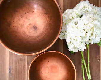 May include: Two copper mixing bowls on a wooden surface. The larger bowl is on the left and the smaller bowl is on the right. A bouquet of white hydrangeas is on the right side of the image.