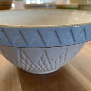 Puede incluir: Primer plano de un cuenco de cerámica blanca con un borde azul claro con un patrón de triángulos repetidos. El cuenco tiene un patrón de diamantes en el exterior y un interior claro. El cuenco está sobre una superficie de madera.