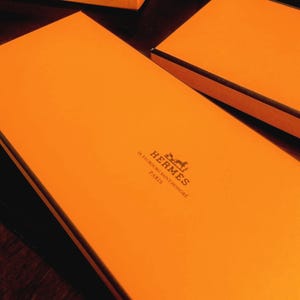 May include: Bright orange boxes with black trim are arranged on a dark brown surface. The boxes have the brand name "HERMES" printed in black, along with the address "24 Faubourg Saint Honore Paris". The boxes are likely for luxury goods.