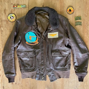 May include: A brown leather bomber jacket with a dark brown fur collar, two front pockets, and a zipper closure. Patches include "Attack Squadron 94" and "Vietnam" text. A "Vietnam Veteran" ribbon and other patches are also visible.