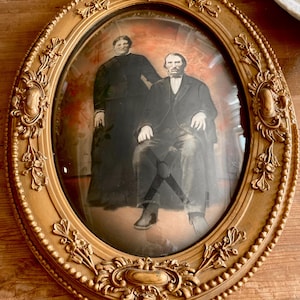May include: An oval-shaped gold picture frame with ornate detailing, featuring a black and white portrait of a couple inside a glass dome.