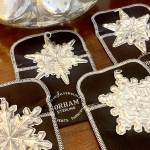 May include: Close-up of several silver snowflake ornaments on black fabric pouches. The ornaments are detailed with intricate designs and hang from small loops. The pouches have white stitching and the text "GORHAM STERLING" is visible.