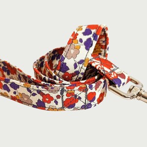 May include: A patterned dog lead with a silver metal clip. The lead displays a repeating floral design in red, purple, and yellow on a cream backdrop. The lead is made of fabric.