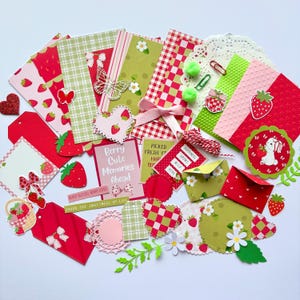 May include: A collection of strawberry-themed paper craft supplies including patterned cardstock, die-cut shapes, tags, envelopes, and stickers. Features include red and green gingham, floral motifs, a pink ribbon, and text reading Berry Cute Memories Ahead.