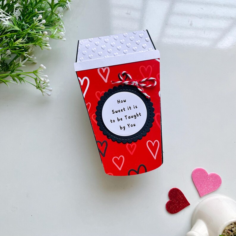 Op de afbeelding: Rode en witte papieren beker met een zwarte cirkel en witte tekst die "How Sweet it is to be Taught by You" luidt. De beker is versierd met witte hartjes op een rode achtergrond.