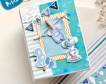 May include: A handmade baby-themed greeting card with a blue and white color scheme. The card features decorative elements like a banner that spells "BABY", baby clothes, an elephant, and the text "Little Prince".