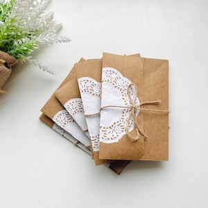 Op de afbeelding: Een stapel bruine papieren enveloppen met witte kanten doilies. De enveloppen zijn samengebonden met touw. De achtergrond is wit, met een kleine plant in een jute pot in de linkerbovenhoek.