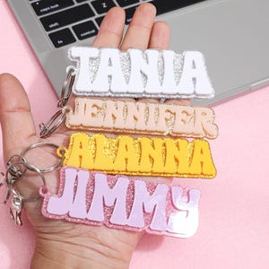 Puede incluir: Cuatro llaveros personalizados con nombres en blanco, melocotón, amarillo y rosa, con detalles brillantes. Cada llavero presenta un nombre diferente en una fuente audaz y retro. Los llaveros se sostienen en una mano, con un teclado de portátil en el fondo.