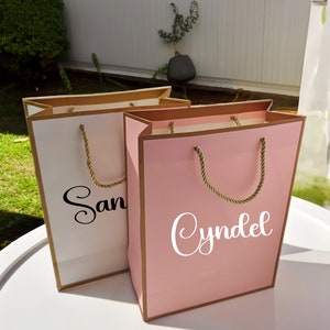 May include: Two gift bags with gold trim. One bag is white with the name "San" written in black. The other bag is pink with the name "Cyndel" written in white.