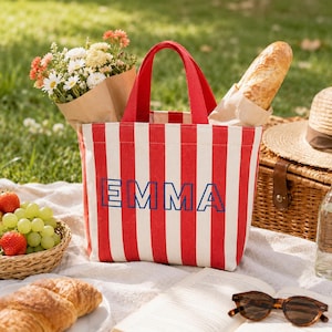 May include: A red and white striped canvas tote bag featuring the name EMMA printed in bold blue capital letters on the front. The bag has two red fabric handles and is filled with a bouquet of white and orange flowers and a long baguette.
