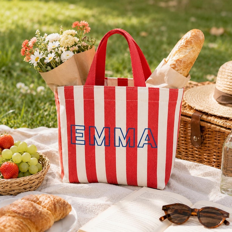 May include: A red and white striped canvas tote bag featuring the name EMMA printed in bold blue capital letters on the front. The bag has two red fabric handles and is filled with a bouquet of white and orange flowers and a long baguette.