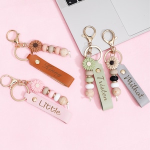 Peut inclure: Porte-clés avec des attaches dorées, des lanières en cuir et des perles décoratives. Les lanières sont de différentes couleurs, dont le marron, le rose, le vert et le gris, avec des noms tels que "Kinny", "Little", "Tristen" et "Mithat" imprimés. Chaque porte-clés a un charme de fleur.
