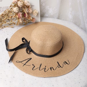 Könnte beinhalten: Ein beigefarbener Sonnenhut aus Stroh mit breiter Krempe und einem schwarzen Band, das zu einer Schleife gebunden ist. Der Name "Arlinda" ist in Schreibschrift auf den Hutrand geschrieben. Ein stilvolles Accessoire.