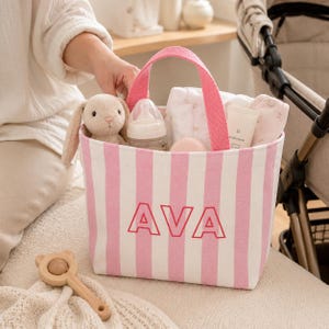May include: A pink and white striped canvas tote bag featuring the name AVA in bold pink letters on the front. The bag contains a plush rabbit toy, a baby bottle, a white cloth, and a tube of cream, and has two pink fabric handles for carrying.