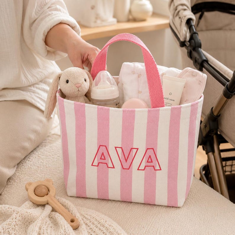 May include: A pink and white striped canvas tote bag featuring the name AVA in bold pink letters on the front. The bag contains a plush rabbit toy, a baby bottle, a white cloth, and a tube of cream, and has two pink fabric handles for carrying.