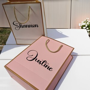 May include: Two gift bags, one white with gold trim and the name "Shannon" printed on it, and one pink with gold trim and the name "Justine" printed on it.