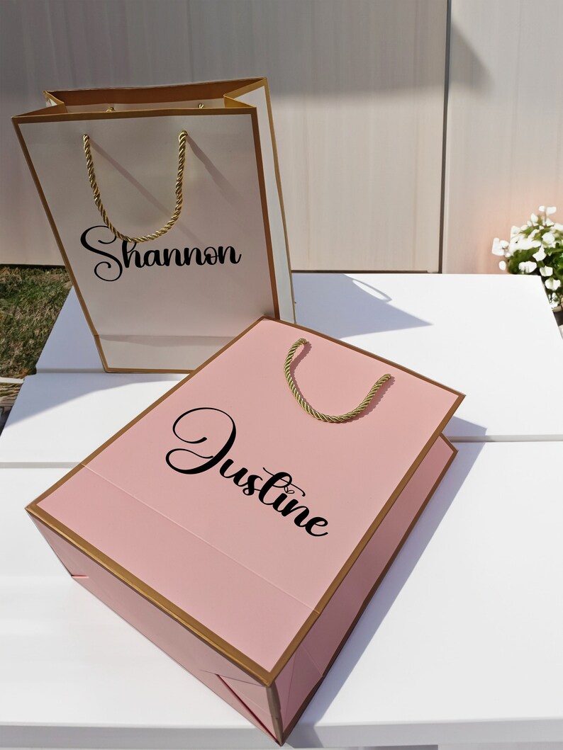 May include: Two gift bags, one white with gold trim and the name "Shannon" printed on it, and one pink with gold trim and the name "Justine" printed on it.