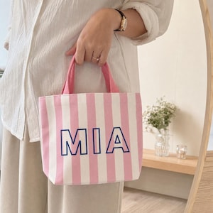 May include: A small canvas tote bag featuring vertical light pink and white stripes. The front of the bag displays the name MIA in bold, dark blue capital letters with a thin outline. It includes two pink fabric handles at the top.