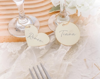 May include: Two place cards with names "Renee" and "Tiana" written on them. The place cards are attached to wine glasses with gold wire.