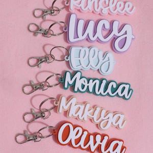May include: Keyrings with names: Kinslee, Lucy, Elly, Monica, Makuya, and Olivia. Each keyring features a name in a layered design with a contrasting colour. They have silver-coloured clasps and key rings, set against a pink background.