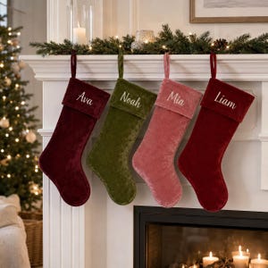 May include: Four velvet Christmas stockings hanging in a row, each featuring a folded cuff embroidered with a name. The stockings are coloured deep red, olive green, dusty rose, and bright red, with the names Ava, Noah, Mia, and Liam stitched in white.
