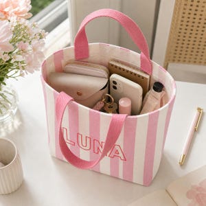 May include: A canvas tote bag featuring vertical pink and white stripes with the name LUNA embroidered in red on the front. The bag has two pink fabric handles and contains a beige wallet, a smartphone, a notebook, and a small bottle.