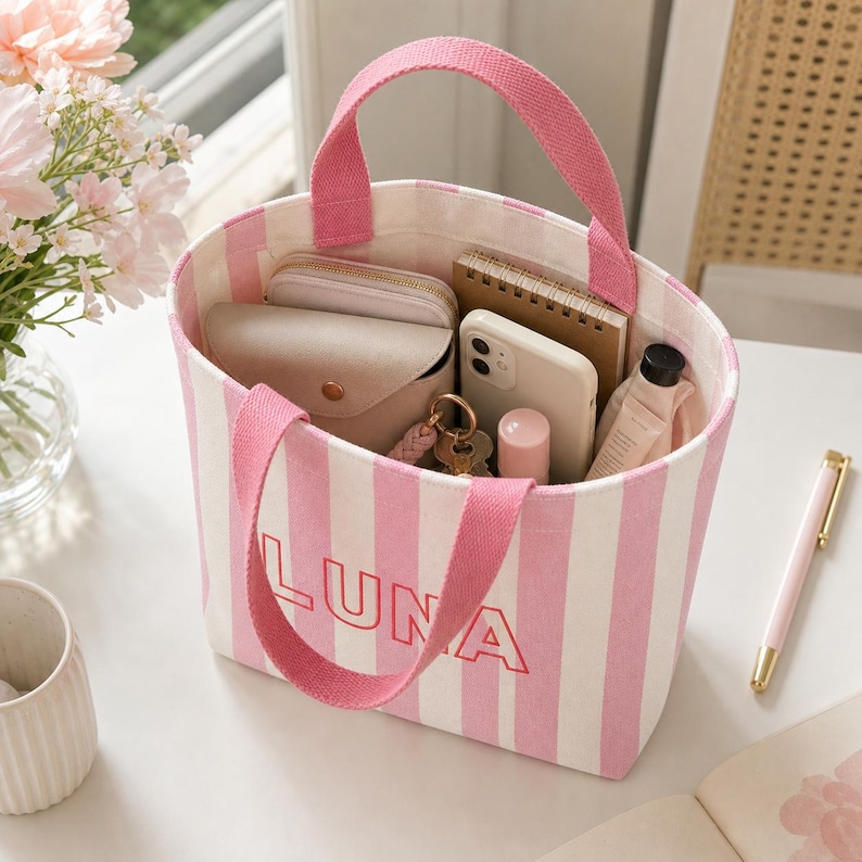 May include: A canvas tote bag featuring vertical pink and white stripes with the name LUNA embroidered in red on the front. The bag has two pink fabric handles and contains a beige wallet, a smartphone, a notebook, and a small bottle.
