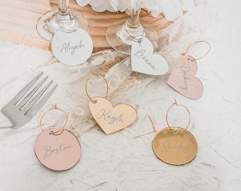 May include: Five personalized acrylic wine glass charms. Two are gold heart-shaped charms with the names "Kayla" and "Leianna" written in white. Two are pink round charms with the names "Boston" and "Darling" written in white. One is a gold round charm with the name "Sandra" written in white.
