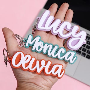 May include: Three personalised name keyrings are displayed. The names are "Lucy", "Monica", and "Olivia", each in a different colour: purple, teal, and orange. The keyrings have a white outline and a silver keyring.