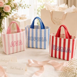 May include: Three small canvas tote bags with vertical stripes and handles. The pink bag features the name AVA, the blue bag features the name SOPHIA, and the red bag features the name EMMA. Each bag has a structured rectangular shape and open top.