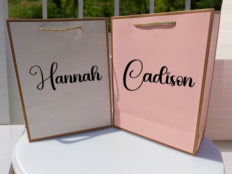 May include: Two gift bags with gold handles and gold trim. The bag on the left is white with the name "Hannah" written in black cursive. The bag on the right is pink with the name "Cadison" written in black cursive.