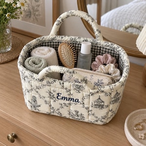 Peut inclure: Un panier de rangement en tissu matelassé avec un motif toile de jouy crème et noir, portant le prénom Emma brodé en bleu sur le devant. L'intérieur est doublé d'un tissu à carreaux vichy noir et blanc, et le panier possède deux anses.