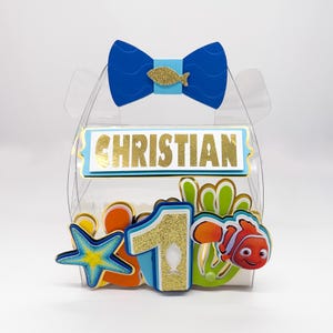 Peut inclure: Une boîte cadeau transparente décorée d'un nœud bleu, d'un poisson doré et du nom "CHRISTIAN" en lettres dorées. La boîte est ornée du chiffre "1", d'une étoile de mer et d'un poisson de dessin animé.