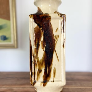 May include: A tall, rectangular ceramic vase with a cream-colored base and dark brown, dripped glaze accents. The vase features a narrow neck, a flared rim, and a flat, square-shaped body with distinct vertical edges and a smooth, glossy finish.