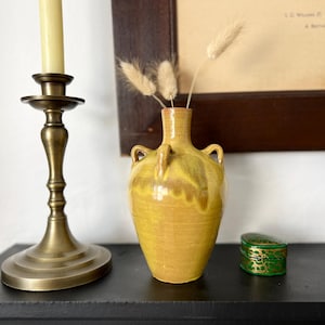 Peut inclure: Un vase en céramique jaune moutarde avec un vernis brillant, un col étroit et deux petites anses latérales. Le vase contient trois tiges botaniques séchées, duveteuses et beige clair. La pièce présente de subtiles variations de couleur.