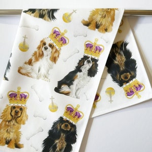 May include: White kitchen towel featuring Cavalier King Charles Spaniels wearing gold crowns with purple accents. The towel also has bone and orb illustrations. The dogs are in various colors, including black, brown, and tan.