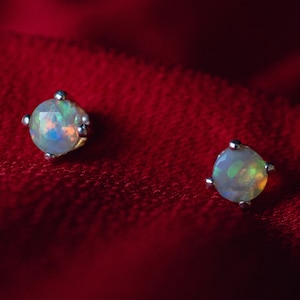 May include: A pair of silver stud earrings with iridescent opal gemstones set in a prong setting. The earrings are on a red velvet background.