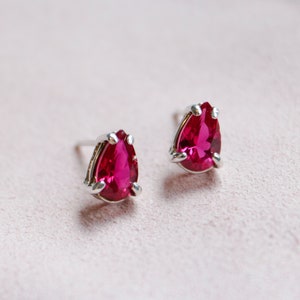 May include: A pair of silver stud earrings with pear-shaped, bright pink gemstones.