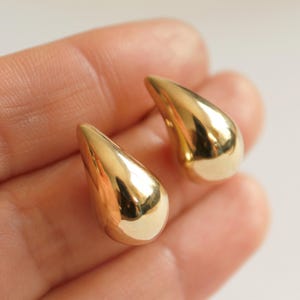 May include: A pair of gold-colored teardrop earrings. The earrings have a smooth, polished surface and a rounded bottom that tapers to a point. The earrings are held in a hand against a white background.