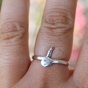 May include: A silver ring with a snake motif. The ring has a polished band and a snake head with two small eye details. The snake's head and tail are raised, giving a 3D effect. The ring is worn on a finger. The ring's design is detailed.