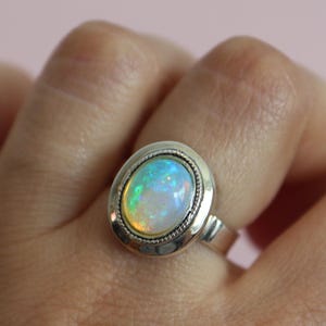 May include: A silver ring with an oval-shaped opal gemstone. The opal has a white base with flashes of green, blue, and yellow.