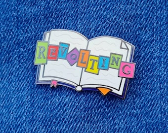 May include: A silver enamel pin shaped like an open book with the word "REVOLTING" spelled out in colorful blocks. The book is white with a blue spine and a yellow bookmark.
