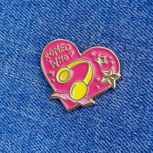 Op de afbeelding: Roze hartvormige emaille pin met de tekst "ROMEO WHO?" en een goudkleurig koptelefoonontwerp. De pin heeft een witte omtrek en kleine witte sterren.
