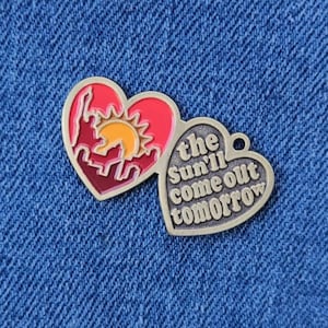 Op de afbeelding: Twee hartvormige metalen pinnen. Eén is rood en roze met een silhouet van een stadsbeeld en een zon. De andere is zilverkleurig met de tekst "the sun'll come out tomorrow".
