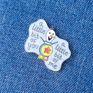 May include: Enamel pin with a smiling snowman design and the text "a little bit of you, a little bit of me" on a light blue shape. The pin has a yellow star and is set against a blue denim background.