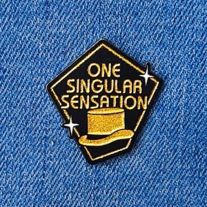 Può includere: Spilla smaltata nera e oro con la scritta "ONE SINGULAR SENSATION" e l'illustrazione di un cappello a cilindro. La spilla a forma di diamante ha uno sfondo nero e dettagli dorati. Due stelle bianche su entrambi i lati.