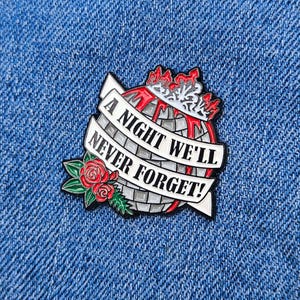 Pode incluir: Pin de esmalte com um design de globo, uma faixa que diz "A NIGHT WE'LL NEVER FORGET!" e uma rosa vermelha. O globo é cinza e branco, com uma coroa e detalhes em vermelho. O pin está sobre um fundo de ganga azul.