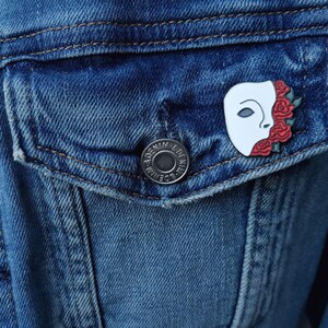 May include: A white enamel pin with a red and white illustration of a face with roses. The pin is attached to a blue denim jacket with a silver button.