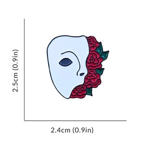 May include: White enamel pin of a face mask with red roses growing out of the side. The mask is 2.5 inches tall and 2.4 inches wide.