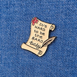 Op de afbeelding: Een gouden emaille pin in de vorm van een rol met een rode waszegel. De rol heeft de tekst "It's hard to be the bard baby" erop.